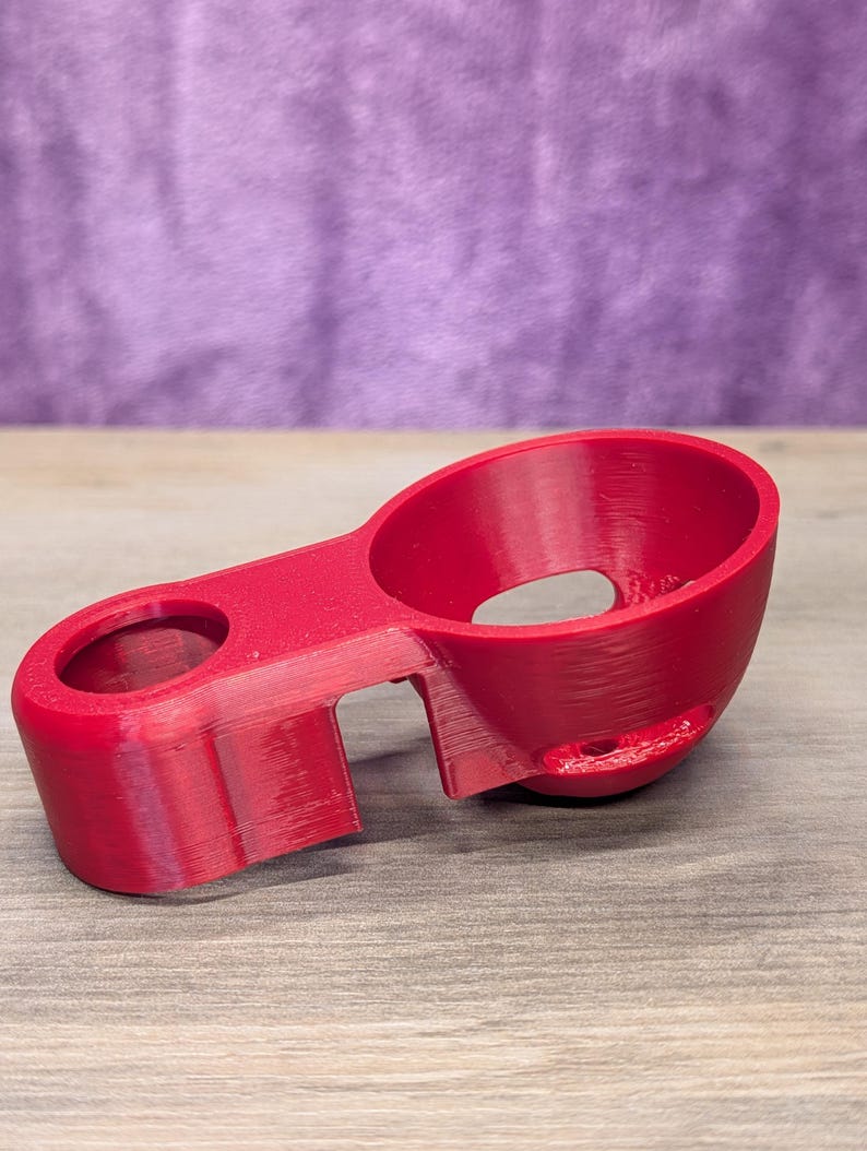 Op de afbeelding: Een helder rood 3D-geprint object met twee cirkelvormige openingen en een verbindingsbrug. Het object heeft een gebogen ontwerp en staat op een houten oppervlak, met een paarse achtergrond.