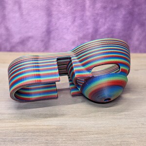 Op de afbeelding: Een 3D-geprint object met een regenboog gestreept patroon. Het object heeft een gebogen, maskerachtige vorm met een opening aan &eacute;&eacute;n kant. De kleuren zijn rood, blauw, groen en geel. Het ligt op een houten oppervlak.