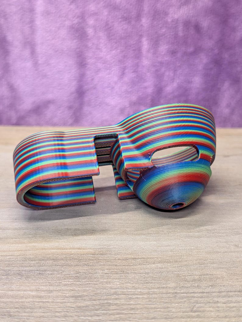 Op de afbeelding: Een 3D-geprint object met een regenboog gestreept patroon. Het object heeft een gebogen, maskerachtige vorm met een opening aan &eacute;&eacute;n kant. De kleuren zijn rood, blauw, groen en geel. Het ligt op een houten oppervlak.
