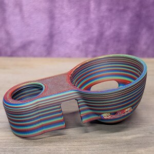 Op de afbeelding: Een veelkleurig 3D-geprint object met een bekerachtige vorm en een handvat. Het object is gemaakt van gelaagd materiaal in tinten rood, blauw, groen en geel. Het heeft een glad, gebogen ontwerp met een gat in het handvat.