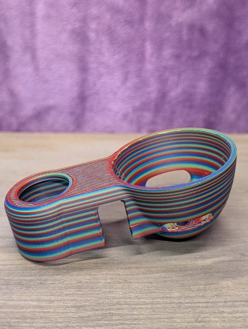 Op de afbeelding: Een veelkleurig 3D-geprint object met een bekerachtige vorm en een handvat. Het object is gemaakt van gelaagd materiaal in tinten rood, blauw, groen en geel. Het heeft een glad, gebogen ontwerp met een gat in het handvat.