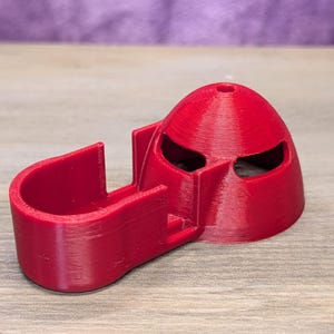 Op de afbeelding: Een rood 3D-geprint object dat lijkt op een helm met oogopeningen en een gebogen, open structuur. Het object is gemaakt van geribbeld plastic en staat op een houten oppervlak, met een paarse achtergrond.