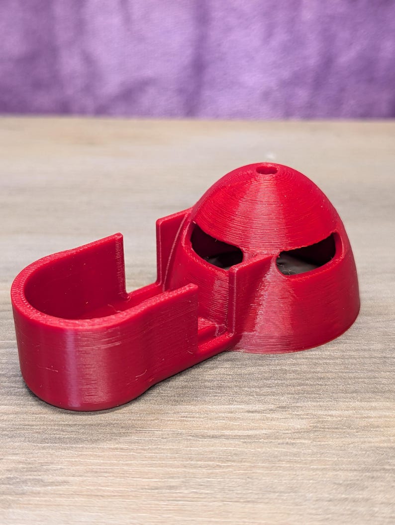 Op de afbeelding: Een rood 3D-geprint object dat lijkt op een helm met oogopeningen en een gebogen, open structuur. Het object is gemaakt van geribbeld plastic en staat op een houten oppervlak, met een paarse achtergrond.