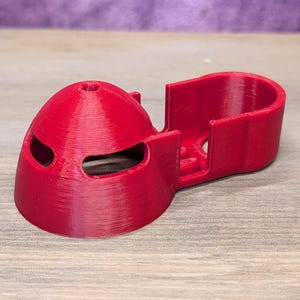 Op de afbeelding: Een helderrode 3D-geprint object, mogelijk een drone-onderdeel, met een afgeronde, helmachtige vorm en twee oogvormige openingen. Het heeft een rechthoekige uitbreiding aan de zijkant. Het object is van plastic en ligt op een houten oppervlak.