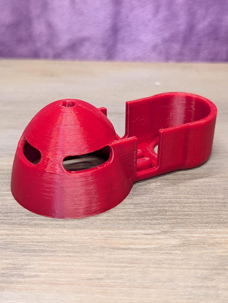 Op de afbeelding: Een helderrode 3D-geprint object, mogelijk een drone-onderdeel, met een afgeronde, helmachtige vorm en twee oogvormige openingen. Het heeft een rechthoekige uitbreiding aan de zijkant. Het object is van plastic en ligt op een houten oppervlak.
