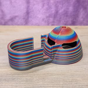 Op de afbeelding: Een 3D-geprint object met een regenboog gestreept ontwerp. Het object heeft een gewelfde bovenkant met twee oogachtige openingen en een gebogen, U-vormige basis. De kleuren zijn rood, oranje, geel, groen, blauw en paars.