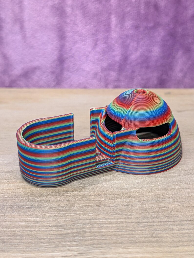 Op de afbeelding: Een 3D-geprint object met een regenboog gestreept ontwerp. Het object heeft een gewelfde bovenkant met twee oogachtige openingen en een gebogen, U-vormige basis. De kleuren zijn rood, oranje, geel, groen, blauw en paars.