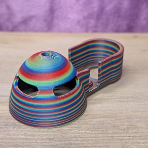 May include: A 3D-printed, multi-colored object with a dome-shaped top and a curved, rectangular base. The object is made of layers of red, blue, green, and yellow. The dome has two eye-like openings.