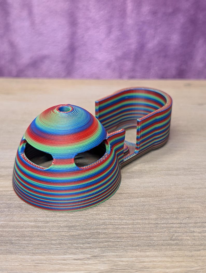 Op de afbeelding: Een 3D-geprint, veelkleurig object met een koepelvormige bovenkant en een gebogen, rechthoekige basis. Het object is gemaakt van lagen rood, blauw, groen en geel. De koepel heeft twee oogachtige openingen.