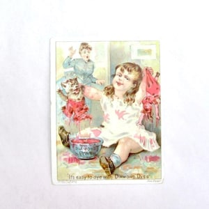 May include: Vintage advertisement for Diamond Dyes. A young girl sits on the floor, smiling, with a bowl of dye and a cat and doll dyed red. The text reads "It's easy to dye with DIAMOND DYES."