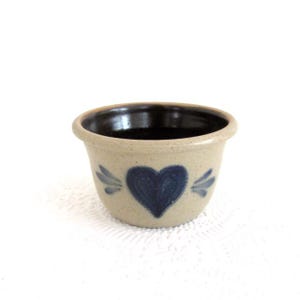 May include: A small, beige stoneware bowl with a dark blue heart design and wing accents. The interior is dark brown. The bowl is likely used for serving or decoration. The bowl is approximately 4 inches in diameter.