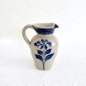 May include: A ceramic pitcher with a gray body and a blue floral design. The pitcher has a curved handle and a spout. The rim and the base of the spout are accented with a dark blue band.