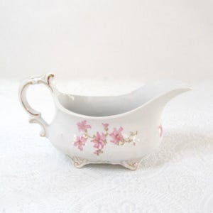May include: White porcelain gravy boat with a delicate pink floral pattern. The gravy boat has a curved handle, a spout, and rests on three small feet. Gold trim accents the handle and feet.