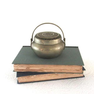 May include: A small, antique metal container with a perforated lid and a handle, resting on a stack of two old books with aged, yellowed pages. The container has a rounded body and a silver finish. The books have dark blue covers.