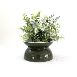 May include: A dark green ceramic vase with a white rim, holding a cluster of green and white artificial eucalyptus leaves. The vase has a unique hourglass shape, adding a touch of modern decor.