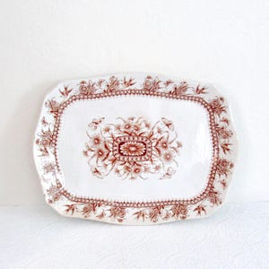 May include: A rectangular ceramic serving platter with scalloped edges, featuring a white base and a dark red transferware pattern. The design includes a central floral bouquet surrounded by a decorative border of flowers, leaves, and butterflies.