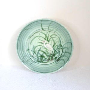 May include: A round, light green ceramic plate with a hand-painted design. The plate features a white bird with a red beak and legs, standing in green reeds. The plate has a ridged edge and is set against a white background.