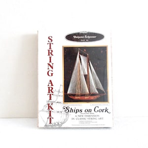 May include: A Bahama Schooner string art kit box featuring a white background with red text reading STRING ART KIT and Ships on Cork. The center displays a finished schooner ship image on a dark cork board with white string and wood hull.