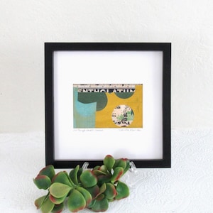 May include: A square black frame containing a mixed media collage artwork. The collage features a yellow background, a light blue shape, dark green circles, and a circular cutout showing vintage text and illustrations, with a ruler visible at the top.