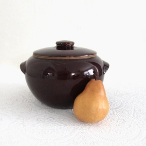 May include: A dark brown ceramic lidded pot with two side handles and a small knob on top. A ripe pear with a light brown skin rests beside the pot. The pot and pear are on a white surface.