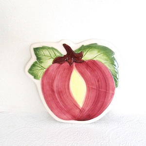 May include: A ceramic decorative plate in the shape of a red fruit with a yellow center, green leaves, and a brown stem. The plate has a white border and is likely used for serving or display.