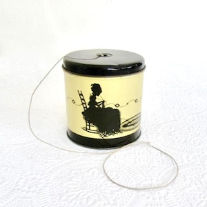 May include: A vintage sewing thread tin with a black lid and base, and a pale yellow body. The body features a black silhouette of a woman knitting. A length of thread extends from the top of the tin, forming a loop.