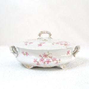 May include: A white porcelain serving dish with a lid, adorned with pink floral patterns. The dish has a decorative handle and gold accents. It is a vintage piece, likely for serving food.
