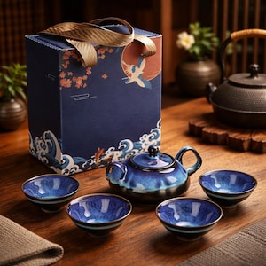 Op de afbeelding: Een blauwe keramische theeset met een theepot en vier bijpassende kopjes, gepresenteerd op een houten oppervlak. De set heeft een glanzende afwerking met een donkerblauwe en lichtblauwe glazuur. Een decoratieve geschenkverpakking met een kraanvogelontwerp is ook aanwezig.