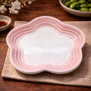 Puede incluir: Un plato de cerámica Le Creuset rosa y blanco, con forma de flor y borde festoneado. El plato tiene un interior blanco y un exterior rosa. El logotipo de Le Creuset está grabado en el borde. El plato está sobre una servilleta de lino beige.