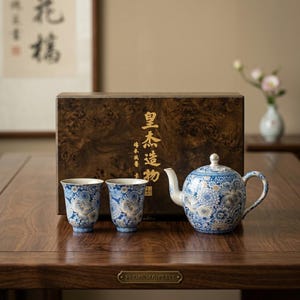 Peut inclure: Service à thé en céramique avec un motif floral bleu et blanc. L'ensemble comprend une théière, deux tasses et une boîte en bois avec des lettres dorées. La table porte une plaque avec l'inscription "Premium Gift Set".