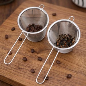 以下が含まれることがあります： 木製の表面に、コーヒー豆と茶葉が入った2つのステンレス製茶こしがあります。各茶こしには長いハンドルと細かいメッシュバスケットが付いています。散らばったコーヒー豆も確認できます。