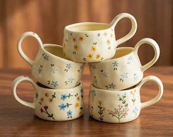 Puede incluir: Seis tazas de cerámica blanquecina con asas grandes, apiladas sobre una superficie de madera. Cada taza está decorada con un patrón floral diferente en amarillo, azul y rojo.