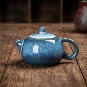 Könnte beinhalten: Eine blaue Keramik-Teekanne mit Deckel und Griff. Die Teekanne hat eine glänzende Oberfläche mit vertikalen Streifen und einen kleinen Knopf auf dem Deckel. Die Teekanne steht auf einer Holzoberfläche.
