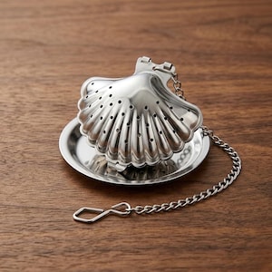 Può includere: Infusore per tè a forma di conchiglia in acciaio inossidabile con vassoio antigoccia abbinato. L'infusore ha un design a cerniera e una catena per una facile rimozione. Posizionato su una superficie di legno.