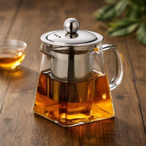 Pode incluir: Uma chaleira de vidro transparente com infusor e tampa de aço inoxidável. A chaleira está cheia de chá de cor âmbar. A alça é curva e a chaleira está sobre uma superfície de madeira. Uma pequena chávena de vidro é visível no fundo.