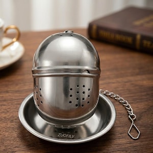 Pode incluir: Um infusor de chá em aço inoxidável com um design esférico, apresentando uma seção inferior perfurada e uma tampa articulada. Inclui um pires pequeno e uma corrente com um gancho. A marca "ZUOFILY" está impressa no pires.
