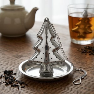 Peut inclure: Infuseur à thé en forme de sapin de Noël en acier inoxydable, avec une chaîne et une petite soucoupe. L'infuseur est posé sur une surface en bois, à côté d'une théière et d'un verre de thé. Des feuilles de thé en vrac sont éparpillées.
