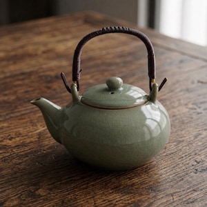 Può includere: Una teiera in ceramica verde celadon con corpo arrotondato e coperchio abbinato. La teiera ha un manico intrecciato marrone e un beccuccio. La teiera è appoggiata su una superficie di legno.