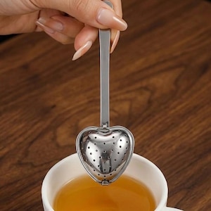 Peut inclure: Infuseur à thé en forme de cœur en acier inoxydable, avec une longue poignée, au-dessus d'une tasse blanche remplie de thé. L'infuseur comporte de petits trous pour infuser les feuilles de thé en vrac. Le thé est ambré.