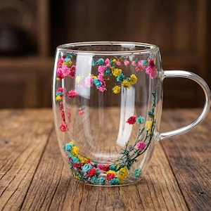 Peut inclure: Une tasse en verre transparent à double paroi, ornée de fleurs séchées colorées incrustées dans le verre. Les fleurs sont roses, jaunes, bleues et rouges, créant un motif floral vibrant. La tasse a une anse argentée et repose sur une surface en bois.