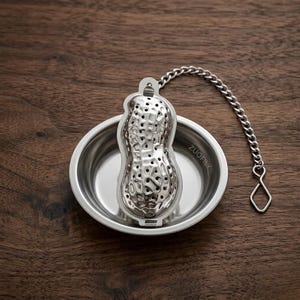 May include: A stainless steel peanut-shaped tea infuser with a chain and a small bowl. The infuser has many small holes and sits inside a round bowl. The chain is attached to the infuser and has a diamond-shaped end. The text "ZUOFILY" is on the bowl.
