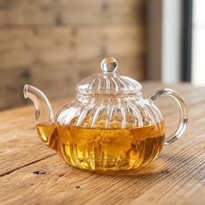 May include: A clear glass teapot with a rounded body and a ribbed design. The teapot is filled with a golden-colored liquid and a yellow flower. It has a curved spout, a handle, and a lid with a small knob. The teapot sits on a wooden surface.
