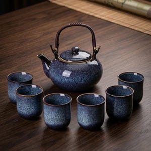 以下が含まれることがあります： 濃い青色の斑点模様の釉薬が施された丸い急須と、暗い色の紐で巻かれた持ち手、そして同じ青と黒の斑点模様で統一された6つの円筒形の湯呑みがセットになった陶器の茶器セット。