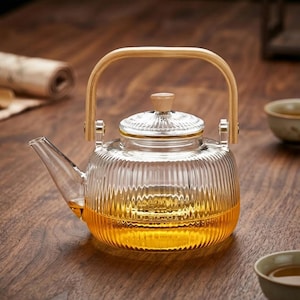 Puede incluir: Una tetera de vidrio transparente con un diseño acanalado, llena de líquido dorado. Cuenta con un asa de madera y una tapa con un pomo de madera. La tetera está sobre una superficie de madera.
