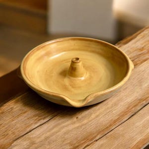 May include: A handmade, ceramic incense holder in a warm, earthy yellow hue. The round dish features a central cone for holding incense sticks and a small spout. The piece is set on a wooden surface.