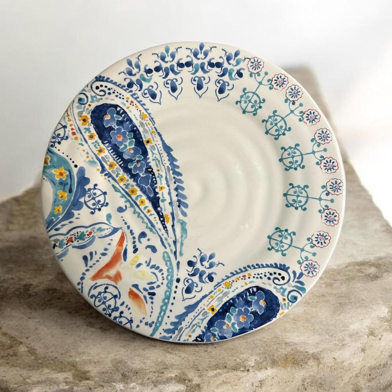 May include: White ceramic plate with a hand-painted paisley design in shades of blue, yellow, and orange. The plate features a central paisley motif and decorative floral border. A unique piece for serving or display.