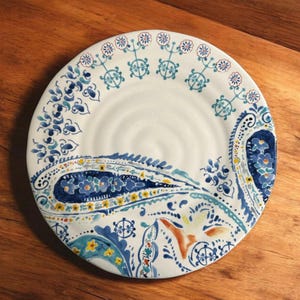 May include: White ceramic plate with a blue and yellow paisley design. The plate features a detailed pattern of floral and geometric shapes in shades of blue, yellow, and orange. The rim has a repeating floral pattern.