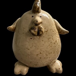 May include: A ceramic chicken figurine with a speckled, off-white glaze. The chicken has a rounded body, small wings, and a prominent beak and wattle. The comb and facial details are a darker brown. The figurine is standing on two feet.