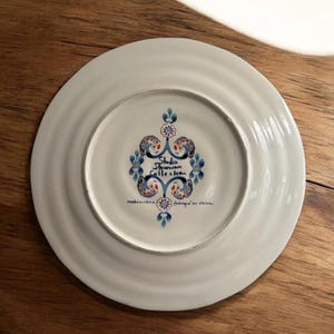 May include: White ceramic plate with a blue, red, and white floral design in the centre. The plate has the text "Studio Donovan Collection" and "made in china" and "fabriqu&eacute; en chine".