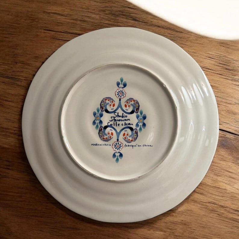May include: White ceramic plate with a blue, red, and white floral design in the centre. The plate has the text "Studio Donovan Collection" and "made in china" and "fabriqu&eacute; en chine".